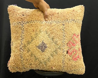 Può includere: Un cuscino quadrato e strutturato con sfondo beige e motivi geometrici rosa, viola e blu. Il cuscino ha un aspetto soffice e tessuto, che suggerisce una consistenza morbida e confortevole. Il cuscino è tenuto da una mano.