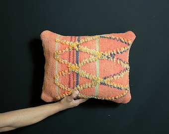 Può includere: Cuscino rettangolare color pesca con un design intrecciato strutturato. Presenta strisce verticali e una serie di accenti ad anello interconnessi, in rilievo, in giallo, arancione e blu. Lo sfondo è nero.