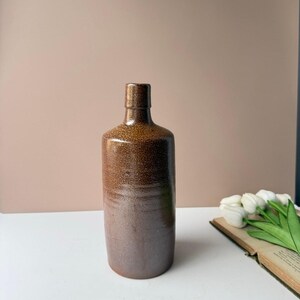 May include: A cylindrical ceramic bottle with a speckled brown glaze. The bottle features a narrow neck and a smooth, rounded rim. The surface texture appears slightly mottled with darker brown tones, transitioning to a solid, darker brown base.
