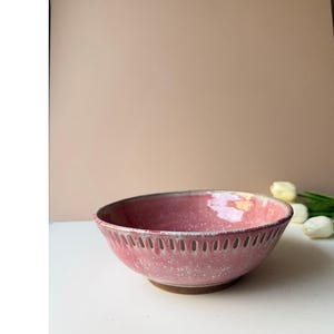 May include: A round ceramic bowl with a speckled pink glaze and a row of vertical cutouts along the upper rim. The interior of the bowl features a glossy pink finish, while the exterior displays a textured, mottled pink and white surface.