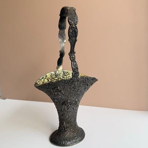 May include: An antique silver-toned basket with an ornate handle and detailed relief carvings. The basket has a flared shape with a decorative rim and a round base. The interior has a gold-colored lining. The basket is displayed on a white surface against a neutral background.