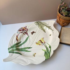 Può includere: Un piatto da portata in ceramica bianca a forma di foglia con un motivo floreale. Il piatto presenta tulipani, felci e farfalle dipinte nei toni del giallo, rosso e verde. I bordi del piatto sono delicatamente smerlati.