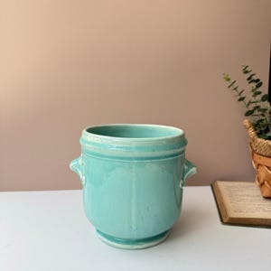 May include: A turquoise ceramic planter with two small handles. The planter has a rounded shape and a slightly flared base. The interior is a lighter shade of turquoise. A small book and a plant in a woven basket are in the background.
