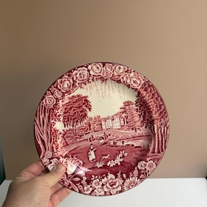 May include: A round ceramic plate featuring a deep red transferware pattern on a cream background. The design depicts a landscape with a castle, a bridge, trees, and figures with a dog, framed by a border of roses and floral motifs.