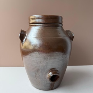 May include: A brown ceramic crock with a lid, two side handles, and a spigot near the base. The glazed surface reflects light, highlighting the vessel's rounded shape. This antique-style container is suitable for storage.