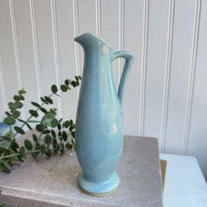 Op de afbeelding: Een kleine, lichtblauwe keramische kan met een rond lichaam en een handvat. De kan staat op een boek met de tekst "The Kinfolk Table".