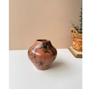 May include: A white ceramic mug with a black handle and the text "But First, Coffee" in a bold, black font. The mug is set against a blurred background, suggesting a cozy setting.