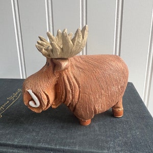 May include: A brown ceramic moose figurine with large antlers and a white accent on its snout. The moose has a textured body and is standing on four small legs. The figurine is placed on a dark surface.