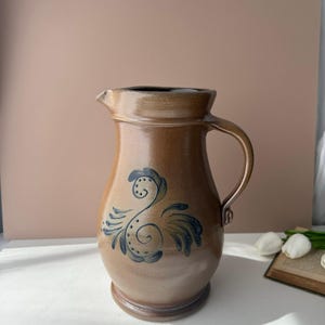 May include: A brown ceramic pitcher with a glossy finish and a curved handle. The body of the pitcher features a dark blue hand-painted floral design with swirling lines and small dots centered on the front. The rim is wide with a small spout.