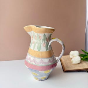 May include: A ceramic pitcher featuring a hand-painted design with a band of pink tulips and green leaves at the top, followed by horizontal stripes in yellow, scalloped pink, solid pink, and a base section with blue diagonal lines and yellow dots.