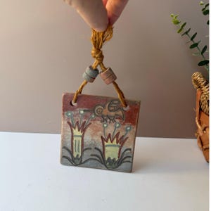 May include: Square ceramic wall hanging with a hand-painted design. The artwork features a bird and stylized flowers in shades of red, brown, blue, and yellow. The piece is suspended by a natural fiber cord with two decorative beads.