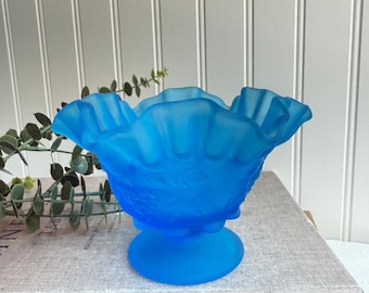 May include: A frosted blue glass bowl with a ruffled rim and a pedestal base. The bowl has a floral design and is sitting on a book titled "Kinfolk Table".