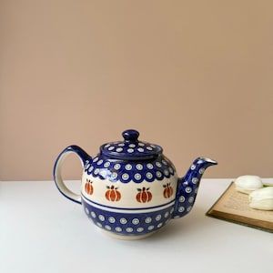 May include: A ceramic teapot featuring a dark blue and cream color palette with a repeating pattern of small blue circles and orange pumpkin illustrations. The teapot has a rounded body, a curved handle, a matching lid, and a tapered spout.