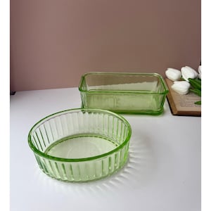 Könnte beinhalten: Zwei Vintage-Servierschalen aus grünem Glas. Eine ist rechteckig, die andere oval. Beide haben ein geripptes Design. Die Schalen stehen auf einer weißen Oberfläche mit rosa Hintergrund. Die Schalen sind leer.