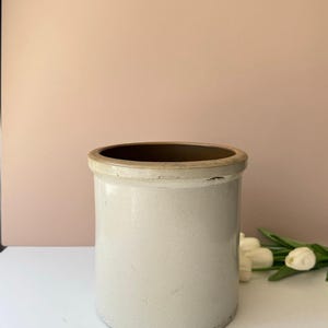 May include: A cylindrical stoneware crock featuring a smooth, light gray exterior finish and a distinct, slightly darker tan-colored rim. The vessel has a wide, open top and a flat base, showcasing a simple, utilitarian design with a glossy glaze.