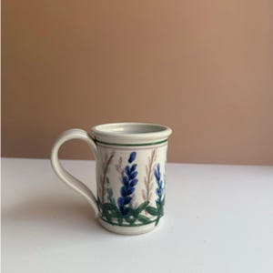 Op de afbeelding: Een keramische mok met een witte basis, met een bloemenontwerp van blauwe en roze bloemen met groene bladeren. De mok heeft een wit handvat en een groene en roze band rond de rand. Geschikt voor koffie of thee.