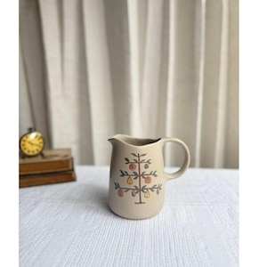 May include: A beige ceramic pitcher featuring a hand-painted illustration of a tree with various colorful fruits including apples and pears. The pitcher has a curved handle and a spout, with a smooth matte finish on the exterior surface.