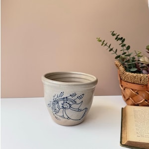 Könnte beinhalten: Beige Keramiktopf mit blauem Engel-Design. Der Topf hat eine runde Form mit leicht ausgestelltem Rand. Der Engel ist mit einem lächelnden Gesicht und Flügeln dargestellt, umgeben von einem kranzartigen Design. Der Topf ist wahrscheinlich für Pflanzen.