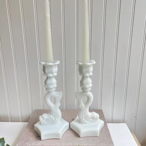 May include: Two white dolphin-shaped candle holders with lit, off-white candles. The candle holders are placed on a stack of books with the text "THE KIN... TABLE". The background is a white paneled wall.