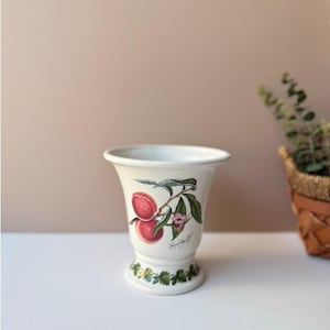 May include: A white ceramic vase with a flared top and pedestal base. The vase is decorated with a colorful illustration of peaches on a branch with green leaves and a pink flower. The base has a green leaf border.