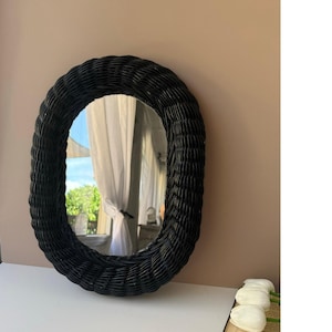 May include: An oval-shaped mirror featuring a thick, black woven wicker frame. The mirror surface is reflective and smooth, surrounded by the dark, textured, and interlaced material of the frame, creating a distinct contrast against the surroundings.