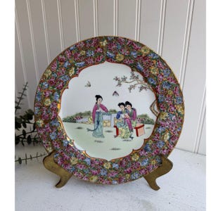 May include: A vintage porcelain plate with a floral border and a scene of three women in a garden. The plate is on a brass stand.