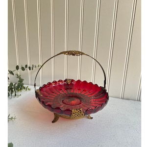 May include: A ruby red glass serving dish with a decorative metal handle and base. The dish has a scalloped edge and a detailed pattern in the center. The metal accents are gold-toned, adding a vintage aesthetic.