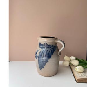 Może przedstawiać: Beżowy dzbanek ceramiczny z zaokrąglonym korpusem i pojedynczym uchwytem, ozdobiony ręcznie malowanym niebieskim wzorem liści z przodu. Wnętrze dzbanka jest ciemne, a zewnętrzna powierzchnia ma nakrapiane, teksturowane wykończenie.