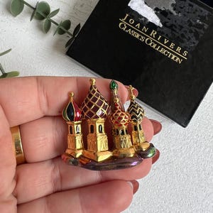 May include: Gold-tone brooch featuring a cluster of five miniature buildings with colorful enamel details. The buildings are reminiscent of Russian architecture and the brooch is labeled "Joan Rivers Classics Collection".