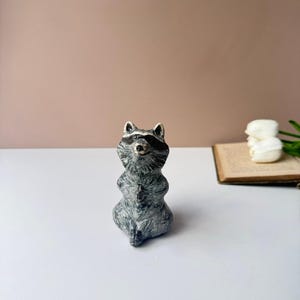 May include: A small, hand-painted ceramic raccoon figurine in a seated position with its front paws pressed together. The raccoon features a grey and white textured coat, a black mask around its eyes, and pointed ears, rendered in a glossy finish.
