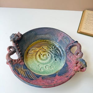 May include: A handcrafted ceramic bowl with a spiral design in the center, featuring shades of green, yellow, and blue. The rim is a mix of red and purple, with dragon-like handles on each side. The bowl has a diameter of approximately 10 inches.
