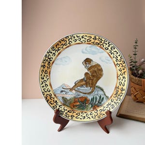 May include: Decorative ceramic plate featuring a monkey illustration. The plate has a cream-colored center with a monkey sitting on a rock, surrounded by a landscape. The rim has a leopard print design. The plate is displayed on a brown stand.
