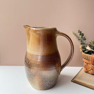 May include: A handmade ceramic pitcher with a large handle and spout. The pitcher has a gradient of colors, transitioning from light tan at the top to a deep brown at the bottom. The speckled texture adds a rustic, artisanal feel. A decorative piece for the kitchen.