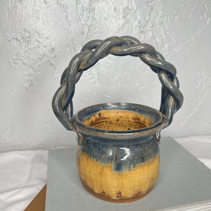 May include: A small, blue and yellow ceramic pot with a braided handle. The pot is sitting on a stack of books.
