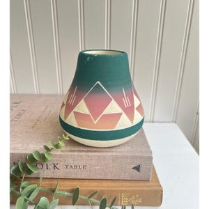 Può includere: Un vaso color verde acqua e crema con disegni geometrici rosa e arancioni. Il vaso ha una base larga e un collo stretto. Il motivo geometrico comprende triangoli e linee. Il vaso è appoggiato su una pila di libri.