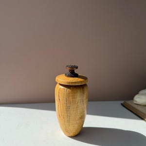 May include: A small, cylindrical wooden vessel with a natural light tan grain and dark vertical streaks. The vessel features a flat, circular wooden lid topped with a small, ornate, dark metallic knob with a textured, circular pattern.