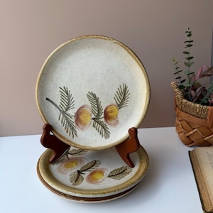 May include: A stack of three ceramic plates with a cream-colored background and a brown rim. Each plate features a hand-painted design of yellow and brown floral elements with green leaves. The plates are displayed on a wooden stand.