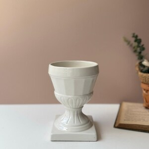 May include: A white ceramic urn-shaped vase with a square base. The vase has a detailed, textured design and a wide opening. The background is a solid, muted pink colour. A book and a small plant are visible in the background.