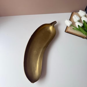 Peut inclure: Un plateau en laiton incurvé et allongé en forme de banane. Le métal présente une finition dorée mate et vieillie avec une surface intérieure lisse et concave, ainsi qu'une tige pointue à une extrémité, offrant un design simple et fonctionnel.