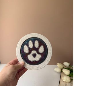 Pode incluir: Um porta-copos circular de cerâmica com um design de pata branca e um pequeno coração no centro. A pata está sobre um fundo escuro, iridescente e rachado, rodeado por uma borda texturizada de cor branco-sujo com linhas radiais.