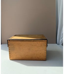 May include: A rectangular wooden box with a natural light brown finish and visible finger joint construction at the corners. The box features a flat lid secured by a thin, dark metal bar that wraps over the top and down the sides of the container.