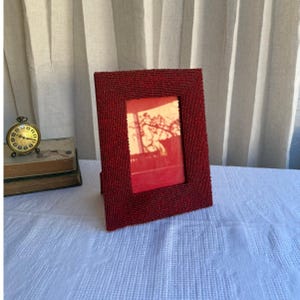 May include: A rectangular picture frame covered in small, deep red beads. The frame features a wide, textured border and a central opening that displays a warm, orange-toned image of a tree silhouette against a bright background.