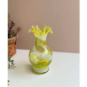 Może przedstawiać: Mały, dekoracyjny szklany wazon z falbanką. Wazon jest w kolorze jasnożółtym z białymi i złotymi wirami. Wazon ma okrągłą podstawę i pękaty korpus.