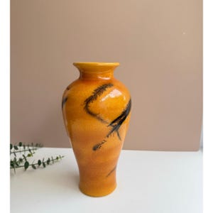 May include: A tall, orange ceramic vase with a glossy finish. The vase features black, feather-like designs that curve around the body. The vase has a rounded base and a slightly flared rim. The background is a neutral tan color.