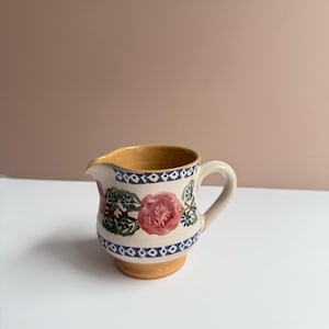 Op de afbeelding: Een kleine keramische creamer met een bloemenontwerp. De creamer heeft een roze roos en groene bladeren, met blauwe en witte geometrische patronen rond de rand en de basis. De handgreep en de binnenkant zijn lichtbeige.
