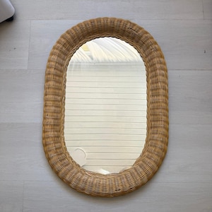 May include: An oval-shaped wall mirror featuring a wide, woven light brown wicker frame. The reflective glass surface is clear and smooth, capturing a reflection of horizontal white slats. The overall design is simple, functional, and symmetrical.