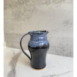 May include: A black and white ceramic pitcher with a flared lip and a handle. The pitcher has a speckled glaze with a dark blue base.