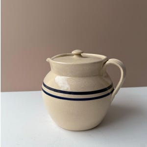 May include: Cream-colored ceramic pitcher with a lid and handle. The pitcher features two dark blue horizontal stripes around the body. The lid has a small knob for easy removal. A classic kitchenware item.