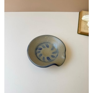 May include: A round, textured ceramic spoon rest with a speckled beige and olive green finish. The center features a circular pattern of dark blue brushstrokes arranged in a radial design, with a small indentation on one side of the rim.