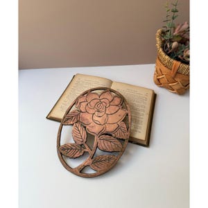 May include: Oval copper-colored metal wall art featuring a detailed rose design. The rose is surrounded by a frame and leaves. The art piece is displayed on a white surface with an open book and a small woven basket in the background.